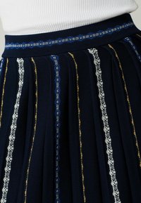 Gonna plissettata blu navy con strisce verticali a contrasto in bianco, blu e oro. Presenta una trama a maglia e una vita aderente.