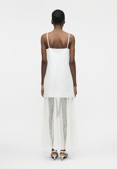 Γυναίκα με σκουρόχρωμο δέρμα και κοντά μαλλιά, φορώντας ένα λευκό μίνιμαλ φόρεμα χωρίς μανίκια με διάφανο δαντελένιο μακρύ φούστα επικάλυψη, όρθια ξυπόλητη μπροστά σε λευκό φόντο.