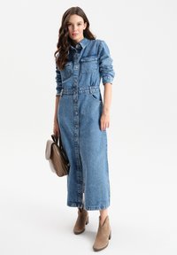 Sukienka koszulowa z denimu w jasnoniebieskim kolorze, z guzikami z przodu, dwiema kieszeniami na piersiach, ściągniętym w talii fasonem i długimi rękawami. Stylizowana z brązowymi botkami do kostek i torebką.