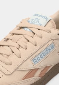 Sneaker in suede beige con lacci crema, linguetta imbottita e suola marrone. Presenta il logo Reebok e sottili accenti marroni.