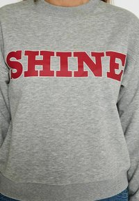 Pilkas sportinis megztinis su įmegztu apykaklės kraštu, puikuojantis dideliu raudonu „SHINE“ grafišku motyvu priekyje. Lygus audinys, laisvas siluetas.