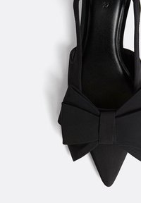 Černá látková bota s špičatou špičkou a velkou mašlí jako akcentem. Design se zavřenou patou, elegantní textura a viditelná hladká podrážka.