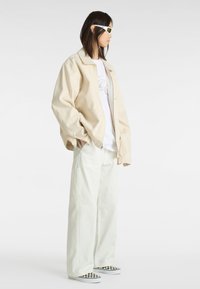 Beige oversized jakke, hvid grafisk t-shirt, lysegrøn brede bukser og sorte og hvide ternede slip-on sko. Hvide solbriller.