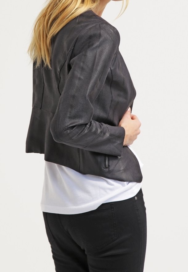 Giacca in pelle nera strutturata con vestibilità su misura, design corto e tasche con zip, indossata sopra una camicia bianca e pantaloni neri.