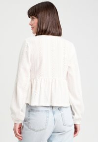 Blouse blanche texturée avec des manches longues et bouffantes, une taille peplum et un délicat détail en œillets sur le tissu. Vue arrière affichée.