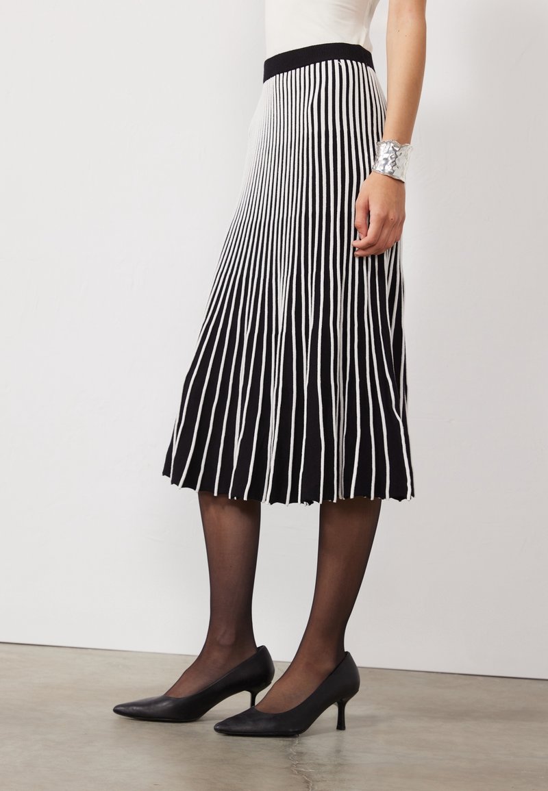 Jupe plissée rayée noir et blanc avec une taille ajustée, en tissu texturé, de longueur genou, associée à des escarpins noirs à bout pointu.