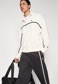 Λευκό μπουφάν με φερμουάρ και μαύρες λεπτομέρειες, σετ με σκούρο παντελόνι που έχει λευκή ρίγα στο πλάι. Το μοντέλο κρατάει μια μαύρη τσάντα duffel.