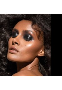 Nærbilde av en models ansikt med mørk, glitrende øyenskygge og definerte kinnbein. Rik, jevn hudtone, krøllete mørkt hår, subtil skjønnhetsflekk.