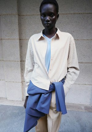 Personne à la peau foncée portant une chemise à carreaux beige clair, un haut bleu, une veste bleue nouée autour de la taille et un pantalon beige, devant un mur en pierre.