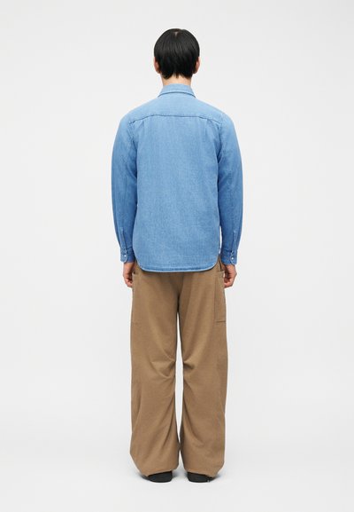 Person standing with back to camera wearing light blue button-up shirt and loose brown pants against plain white background.