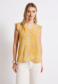 Blusa gialla a fantasia con strisce bianche ondulate, scollatura a V e dettagli a volant sulle spalle. Tessuto leggero, abbinata a pantaloni utility color crema.