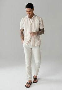 Kortærmet stribet skjorte i beige og hvid, let linnedbukser og brune flip-flops. Modellen står foran en neutral baggrund.