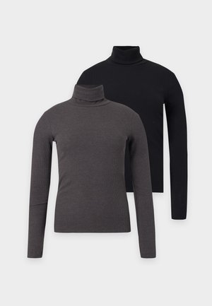 Femme portant un col roulé noir, un jean bleu foncé, des chaussures noires pointues et tenant un sac à main noir sur un fond blanc uni.