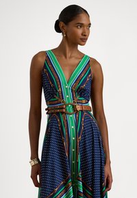 Γυναίκα που φοράει αμάνικο φόρεμα με λαιμόκοψη σε σχήμα V, με εκτύπωση ζώνης και αλυσίδας σε πράσινο, ναυτικό, λευκό και καφέ, διακοσμημένη με χρυσά σκουλαρίκια κρίκους και βραχιόλια.