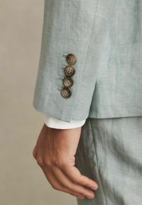 Manecheta unui sacou din in gri deschis cu cinci butoane maro închis, având accente de cusătură albastră și cu o manșetă de cămașă albă care iese în evidență.