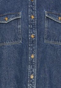 Chemise en denim bleu foncé avec des boutons marron et deux poches à rabat à l'avant, présentant des surpiqûres contrastées le long des coutures et des bords des poches.