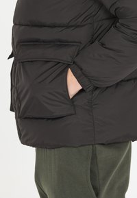 Svart puffjacka med matt finish, med en framficka, elastiska ärmslut och en lätt löst sittande passform. Bärs över gröna tygbyxor.