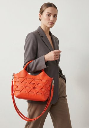 Femme portant un blazer gris et un pantalon marron tenant un sac à main orange texturé avec des poignées courtes et une longue bandoulière.