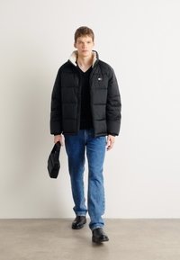 Zwarte pufferjack met crèmekleurige kraag, gecombineerd met een zwart V-hals T-shirt en blauwe jeans. Houdt een zwarte tas vast en draagt zwarte schoenen.