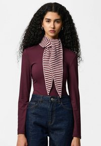 Bordeauxrode top met lange mouwen gecombineerd met donkere denim jeans, voorzien van een gestreepte gebreide sjaal in tinten bordeauxrood en crème die om de nek is gedrapeerd.