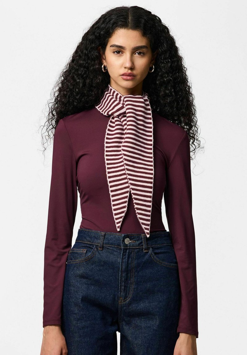 Bordeauxrode top met lange mouwen gecombineerd met donkere denim jeans, voorzien van een gestreepte gebreide sjaal in tinten bordeauxrood en crème die om de nek is gedrapeerd.