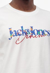 Άτομο που φοράει λευκό T-shirt με πολύχρωμο λογότυπο "Jack & Jones Denim" τυπωμένο στο στήθος.