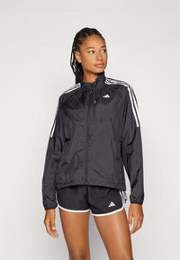 Must musta zip-up jakk, millel on valged külgmised triibud, kõrge krae ja elastsed varrukad, koos mustade lühikeste pükste ja valgete ääristustega.