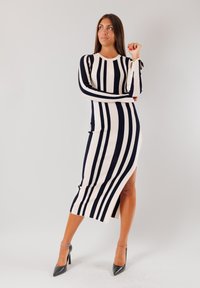 Abito a righe in maglia a costine, caratterizzato da righe verticali blu navy e crema, maniche lunghe e uno spacco alto fino alla coscia. Indossato con tacchi alti neri.
