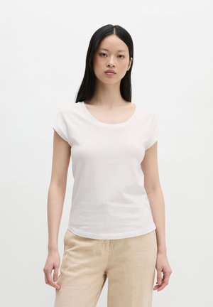Femme aux longs cheveux noirs portant un t-shirt blanc à manches courtes et un pantalon beige, debout devant un fond blanc uni.