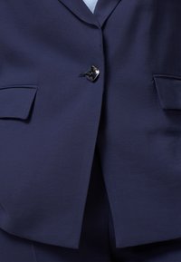 Blazer blu navy con una texture liscia, dotato di un singolo bottone argentato e due tasche a patta frontali. Forma sagomata con colletto classico.
