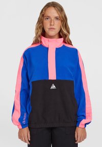 Blå og pink pulloverjakke med høj krave, sort bundpanel, elastiske manchetter og et trekantet logo på brystet. Blød tekstur.