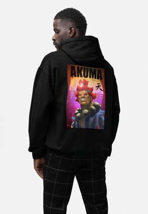 Mężczyzna w czarnej bluzie z kolorową grafiką "AKUMA Street Fighter" przedstawiającą groźną postać o czerwonych włosach i dużych koralach na szyi.