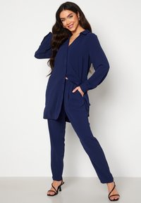 Námořnická modrá, košile s dlouhým rukávem na knoflíky, uvázaná na pase, v kombinaci s odpovídajícími kalhotami. Boty mají tenké pásky a design s vysokým podpatkem.