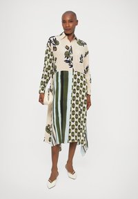 Femme portant une robe à manches longues en patchwork, avec des motifs floraux et géométriques, tenant un petit sac à main blanc et des chaussures blanches à bouts pointus.