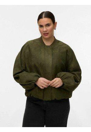 Ženska s temnimi lasmi, oblečena v olivno zeleno bomber jakno z napihnjenimi rokavi in črne hlače, stoji ob preprostem ozadju.