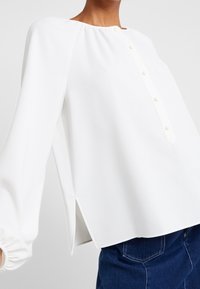 Blusa bianca a maniche lunghe con scollatura tonda e patta con bottoni anteriore, caratterizzata da fessure laterali e polsini arricciati. Tessuto dalla consistenza liscia.
