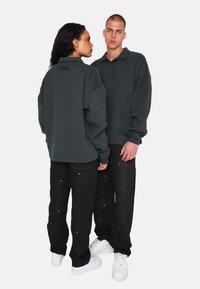 Felpa unisex oversize grigia scura con colletto davanti e dettaglio del logo, abbinata a pantaloni in denim neri e sneakers bianche.