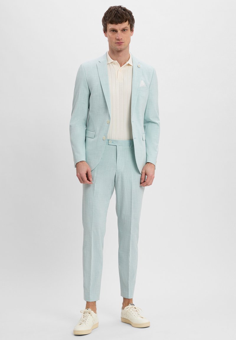 Lichtblauw kostuum met een gestructureerde afwerking, getailleerde jas en bijpassende broek. Draag het over een crèmekleurig poloshirt en combineer het met witte sneakers.