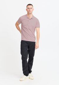 Camisa polo rosa claro con cuello doblado, combinada con jeans ajustados oscuros y zapatillas blancas. El modelo se encuentra de pie con una mano en la cadera.