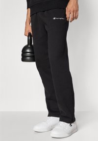 Pantalons de jogging noirs en coton avec ceinture élastique et poches latérales, arborant un logo Champion blanc. Associés à des baskets blanches et à un kettlebell.