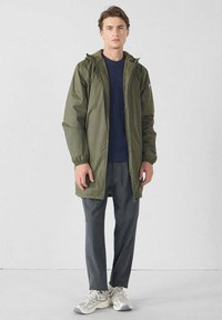 Giacca impermeabile verde oliva con cappuccio, chiusura con zip e polsini elastici. Indossata sopra un maglione di lana blu navy e pantaloni grigi, abbinata a scarpe da ginnastica.