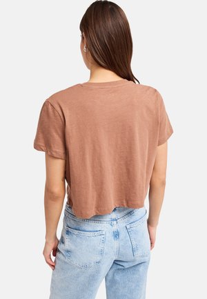 Γυναίκα με ίσια καστανά μαλλιά που φοράει καφέ κοντό T-shirt και ανοιχτό μπλε ψηλόμεσο τζιν, εμφανίζεται από πίσω.