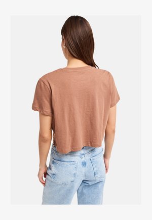 Femme aux cheveux bruns raides portant un t-shirt court marron et un jean taille haute bleu clair, vue de dos.