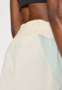 Ανοιχτό μπεζ σορτς με αεριζόμενο πλέγμα και υφασμάτινη λεπτομέρεια σε τόνους του υδάτινου μπλε. Διαθέτει τσέπη με φερμουάρ και οπές εξαερισμού στην πλευρά.