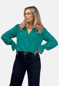 Turkusowa bluzka z głębokim dekoltem w serek i bufkowymi rękawami. Sparowane z ciemnoniebieskimi dżinsami i paskiem z złotą klamrą logo. Gładka faktura materiału.