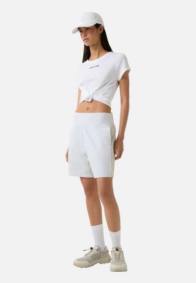 White cotton t-shirt with knotted design, paired with gray shorts and beige sneakers. White cap worn with white ribbed socks.