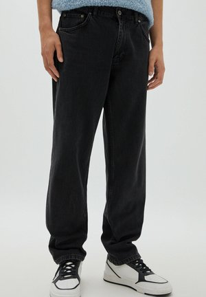 Personne portant un jean noir ample et des baskets blanches avec des détails noirs sur un fond clair uni.