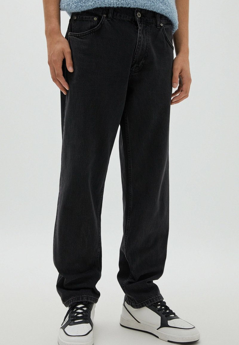 Personne portant un jean noir ample et des baskets blanches avec des détails noirs sur un fond clair uni.