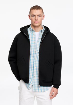 Młody mężczyzna w czarnej bluzie z zamkiem na suwak, noszący pod spodem jasnobłękitną i białą koszulę w kratę oraz białe spodnie, stojący na tle jednolitego tła.