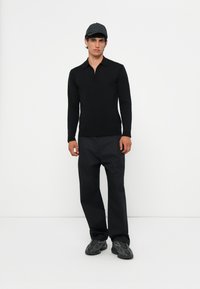 Zwarte longsleeve top met een ritskraag, gecombineerd met losse zwarte broeken en donkere sneakers. Het model draagt een zwarte pet.